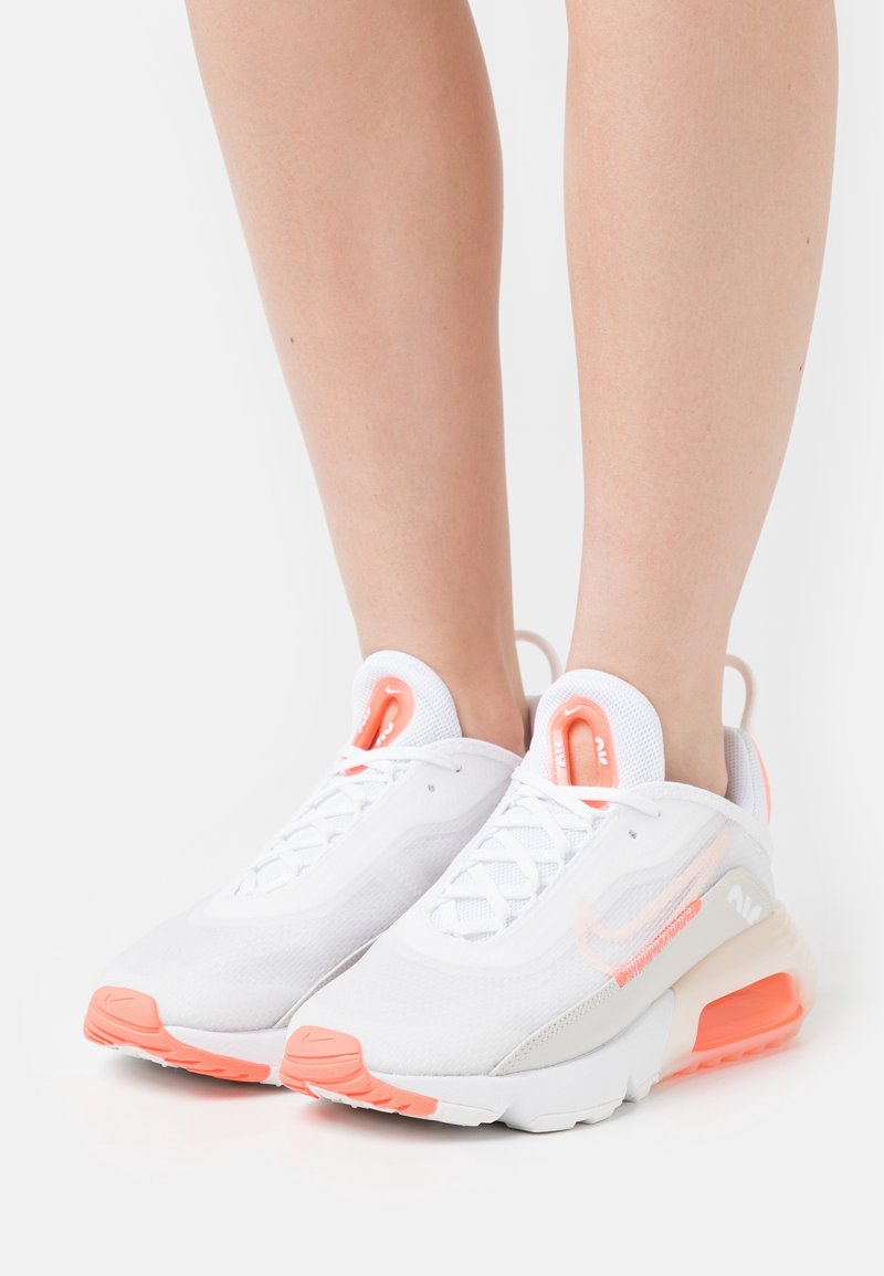 White athletic shoes with orange accents and a mesh upper. Features a rounded toe, laces, and cushioning in the midsole and heel.