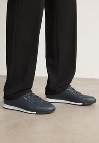 Baskets bleu marine avec une tige en cuir lisse, un design à lacets et une semelle en caoutchouc blanche ; elles présentent un petit logo doré sur le côté.