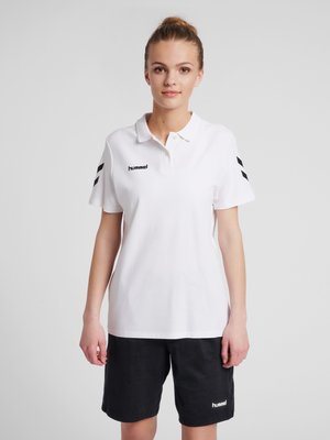 Ung kvinde iført en hvid poloshirt med sorte chevronstriber på ærmerne og sorte shorts, stående mod en ensfarvet baggrund.