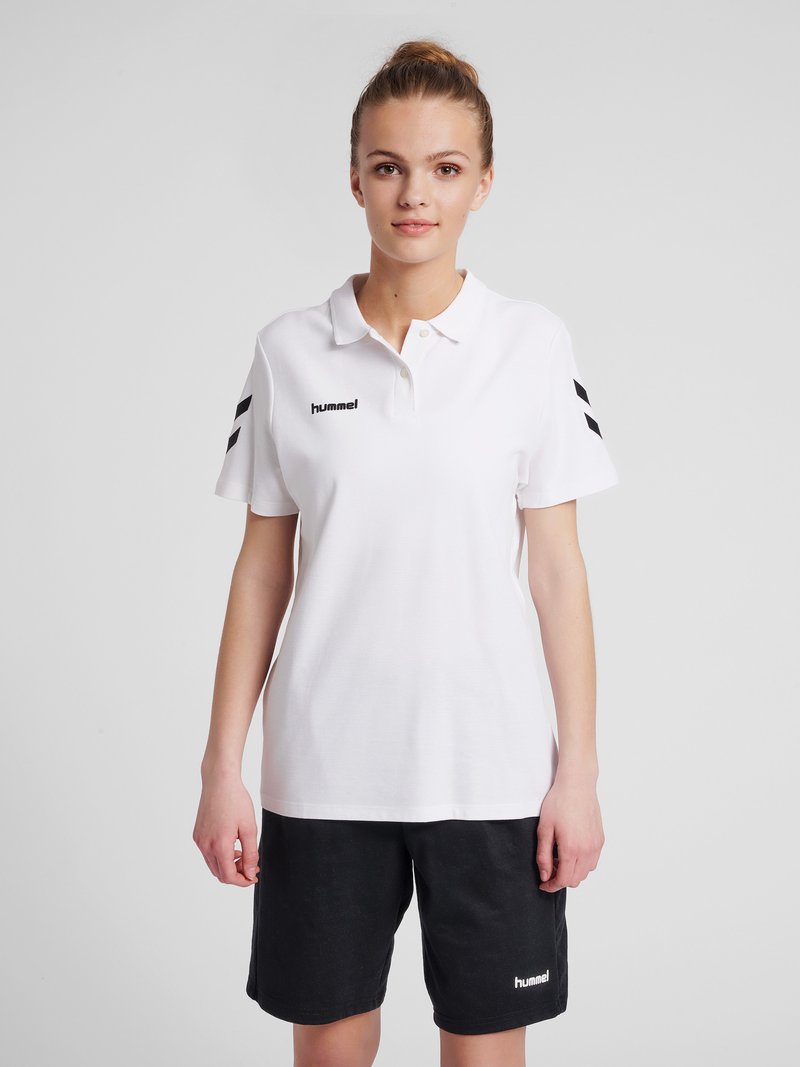 Jeune femme portant un polo blanc avec des bandes en chevron noires sur les manches et un short noir, debout devant un fond uni.