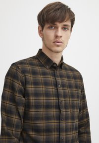 Camisa de franela a cuadros en azul marino oscuro y dorado, con cuello abotonado, bolsillo en el pecho y una textura sutil. Patrón de cuadros clásico.