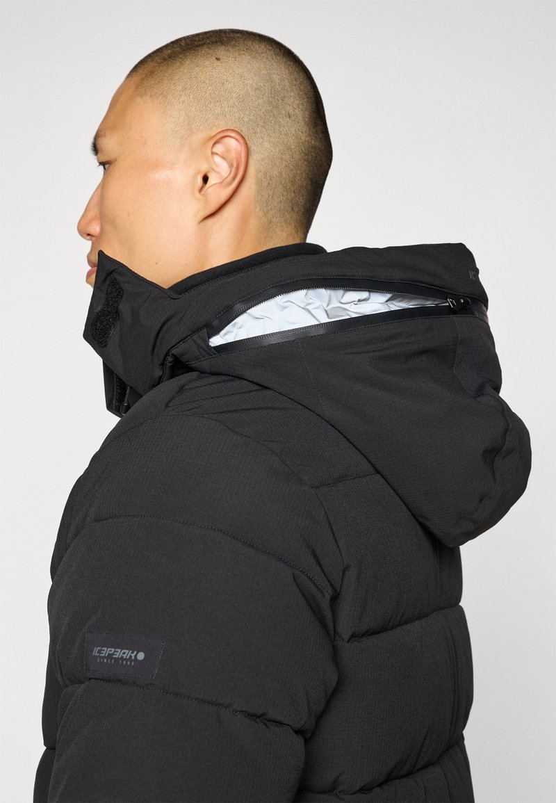 Schwarze isolierte Kapuzenjacke mit strukturiertem Äußeren und glatter Innenauskleidung; verfügt über eine mit einem Reißverschluss verschlossene Tasche auf der Rückseite der Kapuze.