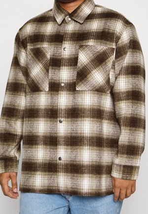 Mann trägt ein braun-creme-kariertes Hemd mit Knöpfen und zwei Brusttaschen, kombiniert mit hellblauen Jeans, vor einem schlichten Hintergrund.