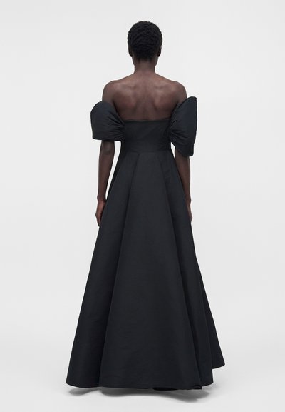 Γυναίκα με κοντά κατσαρά μαλλιά φοράει ένα μακρύ μαύρο φόρεμα με φουσκωτά μανίκια που αφήνουν τους ώμους ακάλυπτους, στέκεται μπροστά σε απλό λευκό φόντο.