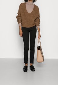 Suéter marrón de punto con un escote en V profundo, debajo un suéter de cuello alto beige, pantalones negros ajustados y zapatos negros de tipo slip-on; sosteniendo un bolso de mano marrón.