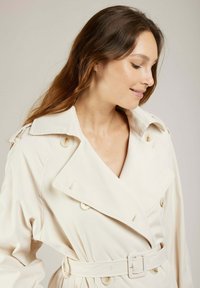 Manteau beige avec un large col, boutons double boutonnage, ceinture à la taille et tissu texturé. Coutures soignées et détails de piqûre subtils.