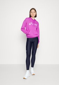 Rosa hettegenser med en hvit logo, kombinert med tettsittende marineblå leggings og hvite sportssko. Outfiten har en avslappet passform og snor i hetta.