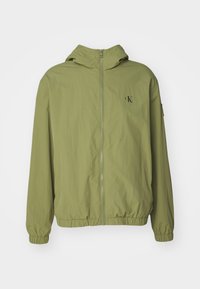 Ljusgrön zip-up jacka med huva, elastiska ärmslut och nederkant, med en liten svart logotyp på bröstet. Slät, lättviktig tyg.