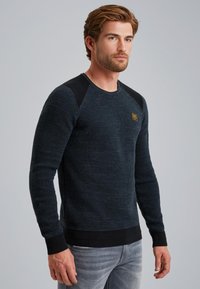 Granatowy sweter z okrągłym dekoltem o teksturowanym splocie, z czarnymi akcentami na ramionach, ściągaczami na rękawach i u dołu, w zestawie z jasnoszarymi dżinsami.