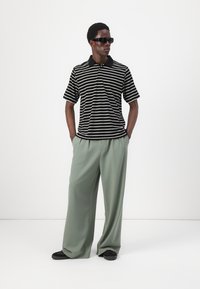 Black and white striped polo shirt with short sleeves and a black collar, paired with loose-fitting, light green trousers and black shoes.