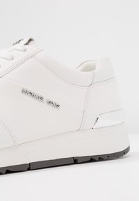 Sneaker bianca in pelle con logo "Michael Kors" argentato, dettagli testurizzati, design senza cuciture e suola nera a contrasto.