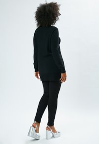 Longo maglione nero con polsini e orlo a coste, abbinato a leggings neri e scarpe col tacco alte argentate, catturato da dietro.