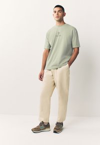 Svetlo zelena majica s logotipom "Guinness", krem hlače in rjavi supergi New Balance z zelenimi poudarki. Minimalistična zasnova.