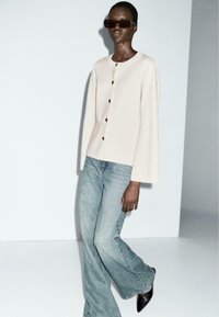 Lichtbeige cardigan met ronde hals en knoopsluiting aan de voorkant, gecombineerd met lichtblauwe flared jeans en zwarte puntige hakken, gedragen met een donkere zonnebril.