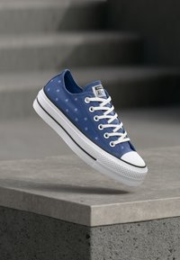 Μπλε αθλητικό παπούτσι Converse με λευκά κορδόνια και σχέδια αστεριών που αιωρείται πάνω από μια τσιμεντένια άκρη με σκάλες στο φόντο.