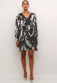 Robe plissée à motifs noir et blanc, à manches longues et en V. Le tissu léger accentue le mouvement. Associée à des talons argentés.