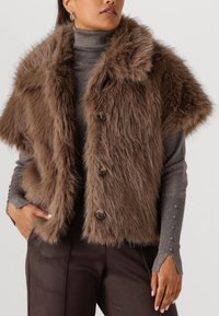 Nep bont vest in bruin met een brede kraag, vier knopen sluiting en zijzakken. Draagt over een grijze coltrui met knoopmanchetten.