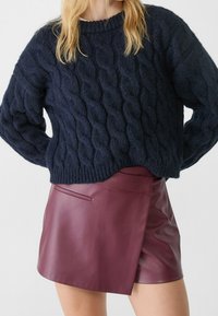 Maglione blu navy a treccia con maniche lunghe e scollo a coste, abbinato a una mini gonna in pelle bordeaux con piega laterale e tasca.