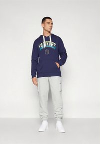 Μπλε μπλούζα με κουκούλα και το κείμενο "Yankees" σε διαβαθμισμένα χρώματα, συνδυασμένη με ανοιχτό γκρι cargo παντελόνι jogger και λευκά αθλητικά παπούτσια.