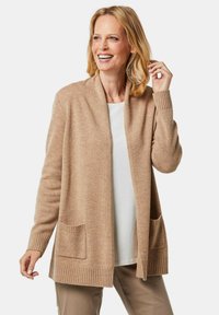 Beige cardigan met een sjawlboord, geribde manchetten en twee voorzakken. Gemaakt van een zachte stof, heeft het een relaxed fit en rechte zoom.