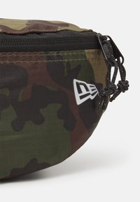 Kamouflage midjeväska med ett hållbart tyg, svart dragkedja och logotypdetalj. Har en rundad form och justerbar dragsnörning på sidan.