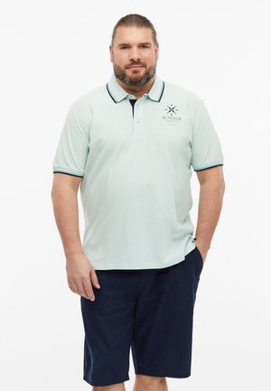 Mężczyzna z brodą w jasnoniebieskiej koszulce polo i ciemnoniebieskich szortach stoi z jedną ręką w kieszeni na białym tle.