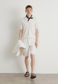 Polo blanc à manches courtes, shorts blancs décontractés, tenant une veste blanche, associé à des sandales à glissière sombres. Fond uni et clair.