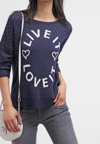 Mörkblå stickad tröja med vita prickar och texten "LEVA DET ÄLSKA DET" i en cirkulär design, med ribbade muddar och nederkant.