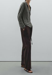 Person wearing a gray pinstripe button-up shirt, loose dark brown pants, and brown slide sandals, standing with hands in pockets.