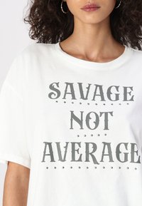 Witte katoenen T-shirt met opvallende grijze tekst die "SAVAGE NOT AVERAGE" leest, versierd met kleine zilveren studs voor detail.