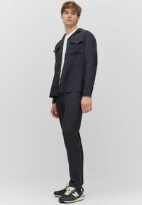 Donkerblauwe jas met knopen, twee borstzakken en bijpassende slim-fit broek; gecombineerd met zwarte sneakers met witte accenten en veters.