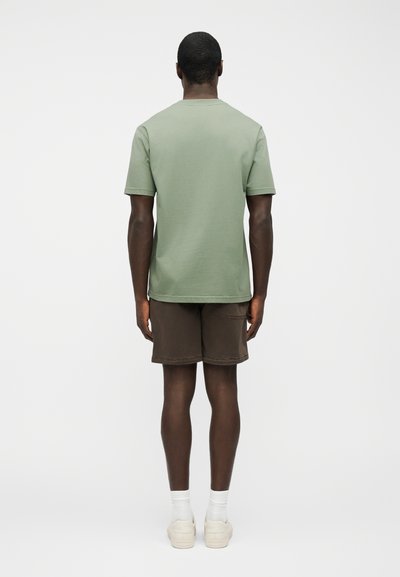 Άνδρας που στέκεται με το πρόσωπο προς τα πίσω, φορώντας ανοιχτό πράσινο κοντομάνικο πουκάμισο, σκούρα καφέ σορτς, άσπρες κάλτσες και εκρού αθλητικά παπούτσια.