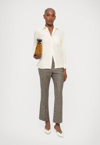 Camisa blanca de manga larga con cuello, pantalones de cuadros en tono beige y tacones blancos de punta. Sosteniendo un bolso de ante marrón.