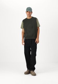 Felpa bicolore verde oliva con motivo a rombi, abbinata a pantaloni cargo neri e scarpe beige, completata da un cappellino teal.