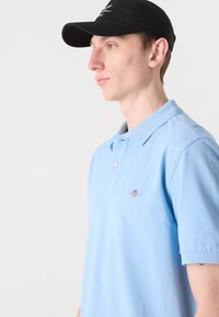 Lyseblå polo-shirt laget av bomull, med en klassisk krage, to knapper og et lite brodert design på brystet.