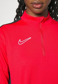 Punainen urheilu toppi, jossa on puoliveto-kaulus, valmistettu kevyestä materiaalista; siinä on valkoinen Nike-logo ja mustia aksentteja hartioilla.