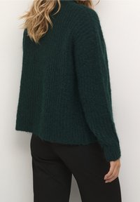 Maglione lavorato a maglia di colore verde scuro, con una finitura testurizzata, vestibilità ampia, maniche lunghe e orlo arrotondato, abbinato a pantaloni neri.