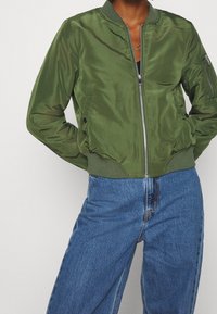Zielona kurtka bomberka o gładkiej, błyszczącej fakturze, z prążkowanymi mankietami i zapięciem na zamek błyskawiczny z przodu. W zestawie z jasnoniebieskimi dżinsami.