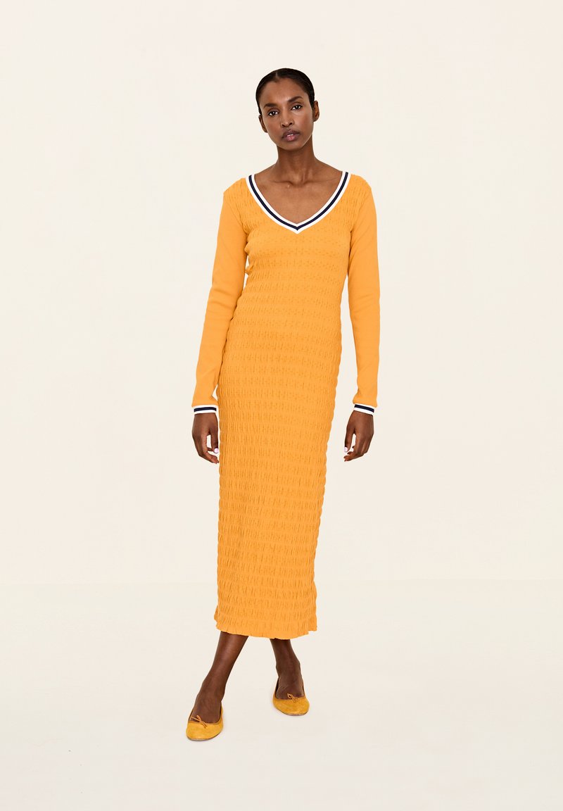 Gelbes Strickkleid mit taillierter Silhouette, V-Ausschnitt und kontrastierendem Marineblau-Besatz am Kragen und an den Ärmeln. Glatte Textur.