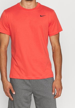 Mężczyzna w koralowym t-shircie Nike z krótkim rękawem i szarych spodniach dresowych, stojący z rękami swobodnie opuszczonymi wzdłuż ciała na jednolitym tle.