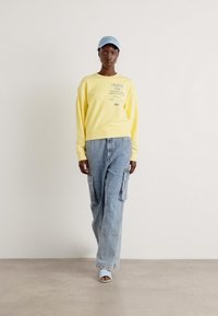 Sweatshirt giallo con testo stampato, abbinato a pantaloni cargo in denim azzurro chiaro con tasche, e cappellino azzurro chiaro e sandali.