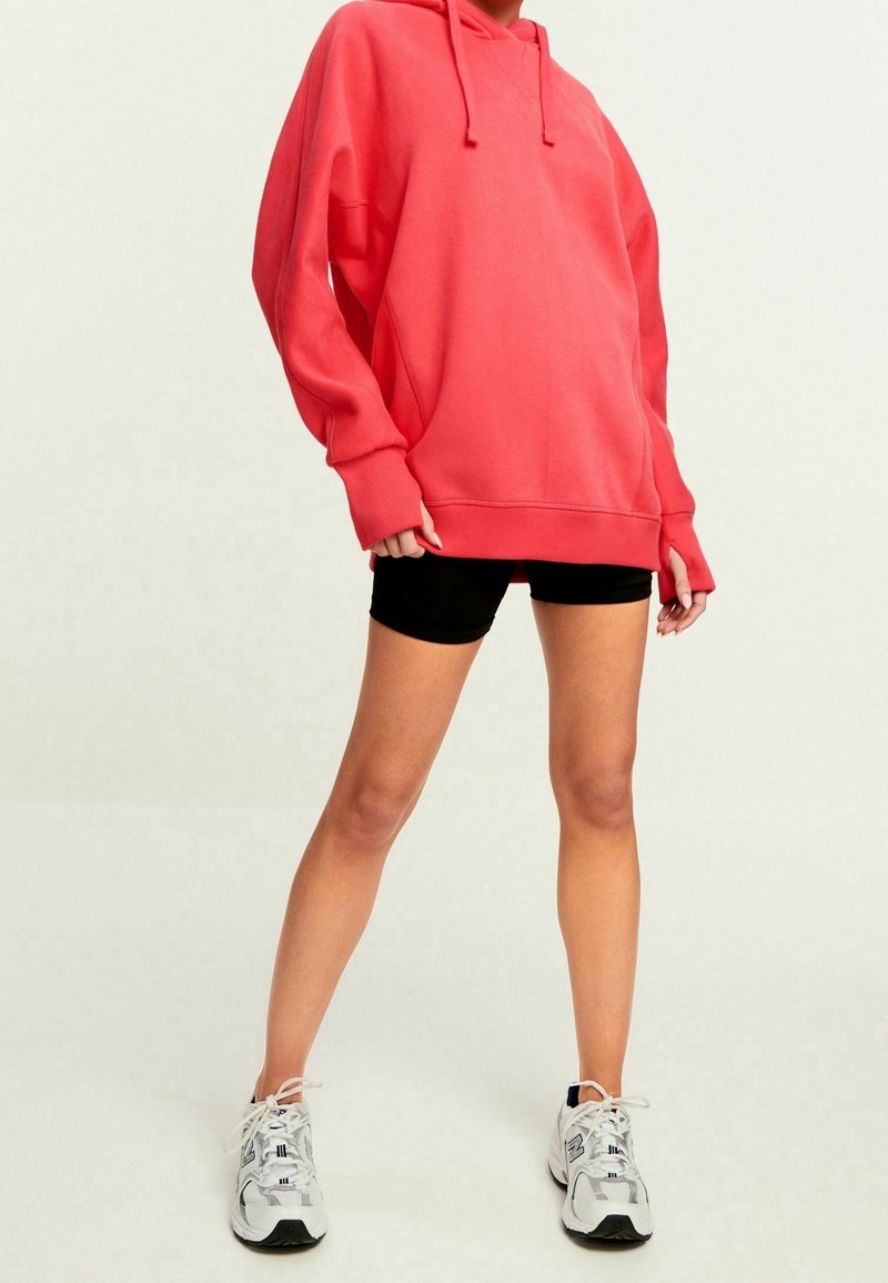 Sweat à capuche surdimensionné corail avec capuche à cordon, manches longues et poignets côtelés. Porté avec un short noir et des baskets blanches.