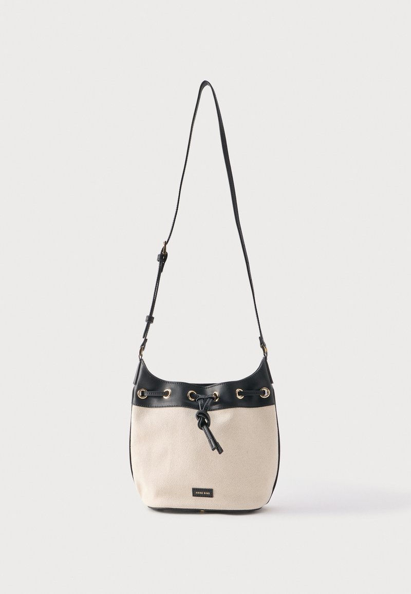 Beige canvas emmer tas met zwarte leren afwerking, verstelbare schouderband, gouden rijggaatjes en vooraan sluiting met trekkoord in knoopvorm.