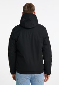 Giacca nera con cappuccio e un esterno liscio e strutturato. Presenta un colletto alto, una vestibilità aderente e un design semplice. Indossata sopra jeans azzurri chiari.