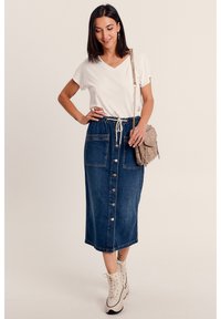 Denimrock mit einer durchgehenden Knopfleiste, zwei großen Taschen, einem elastischen Taillenbund mit Kordelzug, kombiniert mit einem weißen V-Ausschnitt-T-Shirt und beigen Stiefeln.