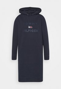 Navy blue hooded dress with a front pocket, featuring the "TOMMY HILFIGER" logo adorned with rhinestones. Made from a cotton blend, it has a relaxed fit and long sleeves.