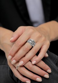 Sølvring i metal med en bølget, skulpturel form sidder på fingrene med lys nude neglelak; hænderne hviler afslappet på en mørk stofoverflade.