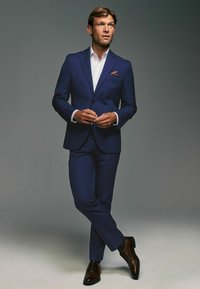 Marineblauer Anzug mit einem einknöpfigen Sakko und schmalen Revers, kombiniert mit einem weißen Hemd. Braune Leder Schuhe und ein Einstecktuch setzen Akzente.