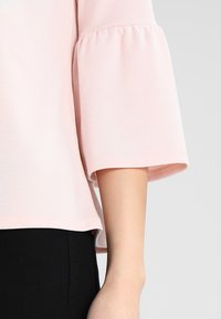 Světle růžová blůza má kulatý výstřih, 3/4 rukávy ve tvaru zvonu s nařasenými detaily a rovný spodní lem. Vyrobena z měkké, hladké látky.