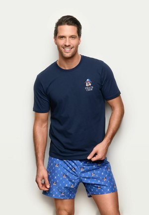 Hombre sonriente con camiseta azul marino "Pirate Crew" y pantalones cortos azules con patrones de anclas y calaveras piratas, de pie frente a un fondo claro.